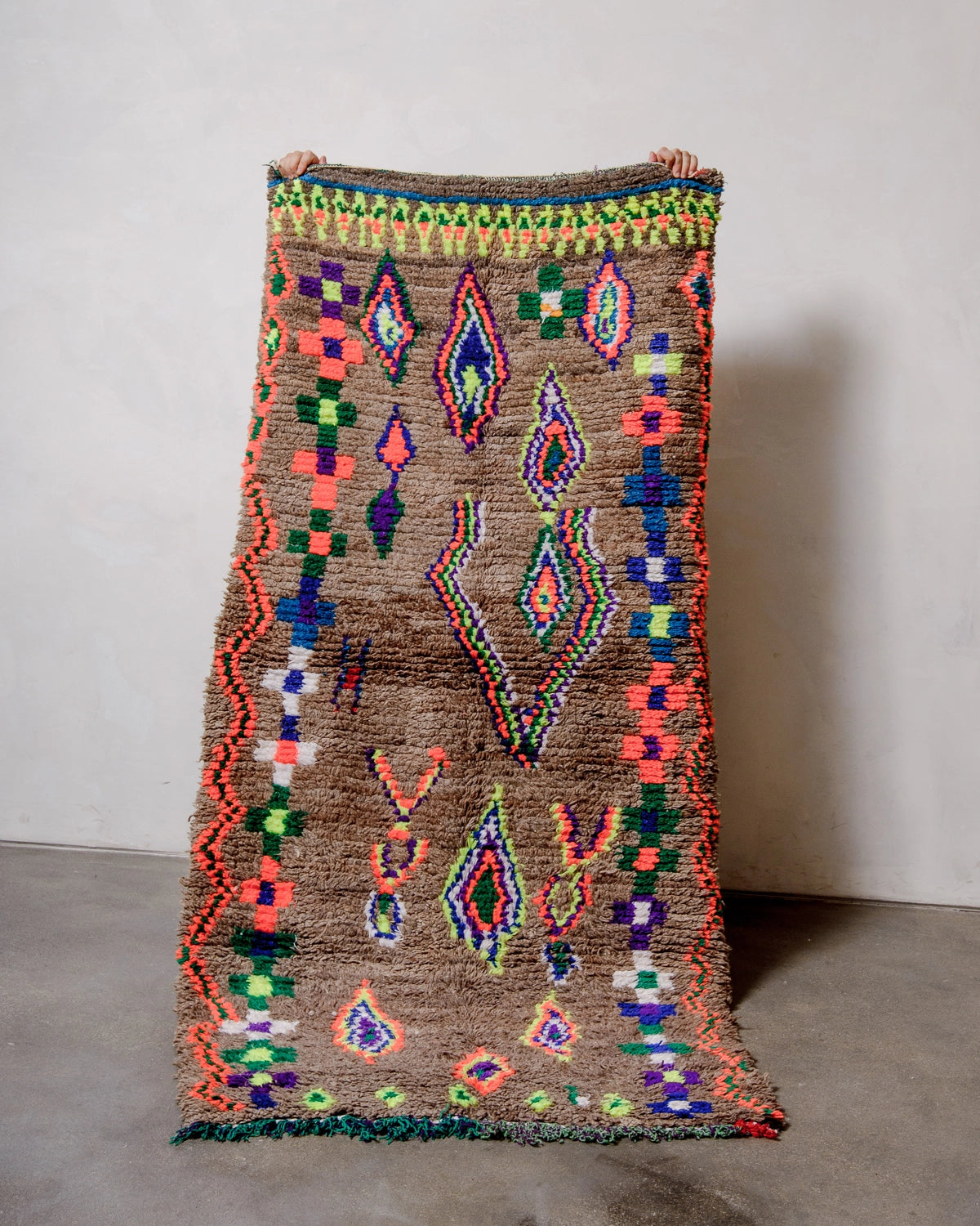 Vintage Berber Rug made of brown wool with design details in neon, purple, blue, green cotton, held up against a white wall.
